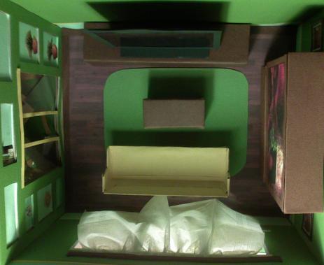 Model kartonowy pokoju: zielone ściany, drewniana podłoga, miniaturowe meble z kartonu, sofa przykryta folią, półki z obrazkami na ścianie.