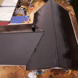 Widok z góry na nowy dach pokryty ciemnymi, łuskowatymi płytkami, z widocznym kominem i częścią rynny, otoczony ziemistym terenem i zielenią.