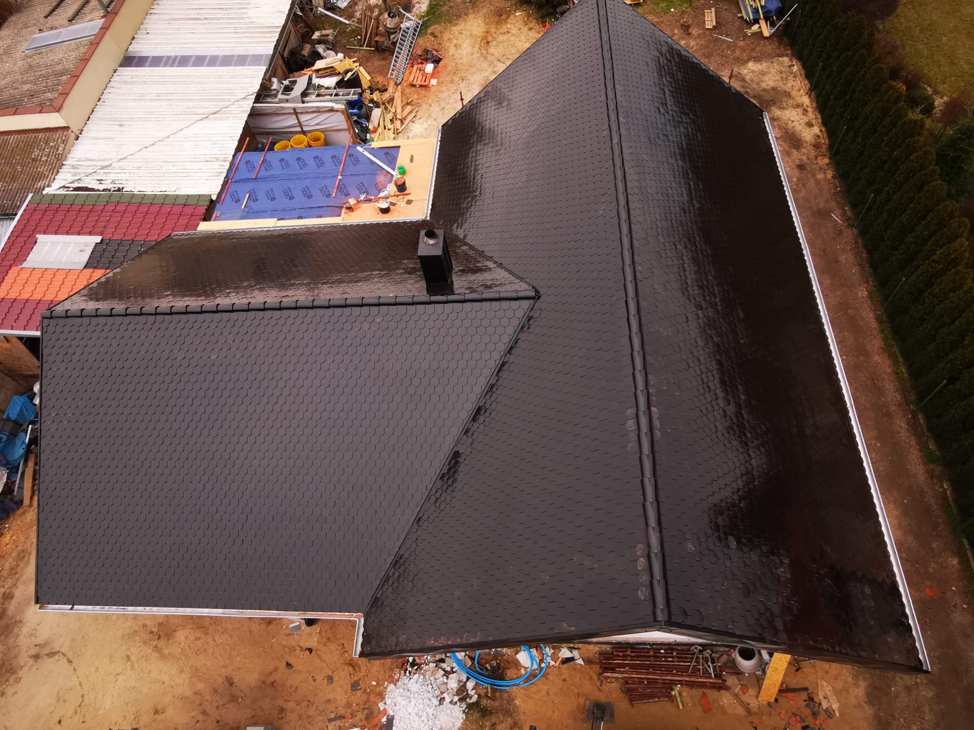 Widok z góry na nowy dach pokryty ciemnymi, łuskowatymi płytkami, z widocznym kominem i częścią rynny, otoczony ziemistym terenem i zielenią.