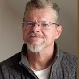 Krzysztof Uchacz-AUTORYTET - Portret mężczyzny w okularach, z siwiejącą brodą i wąsami, ubrany w szary sweter z grubym splotem, na neutralnym tle.