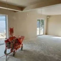 Pomieszczenie w trakcie prac wykończeniowych, widoczna maszyna do nakładania tynku, ściany pokryte świeżym tynkiem, podłoga częściowo zakryta resztkami materiału.