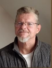 Portret mężczyzny w okularach, z siwiejącą brodą i wąsami, ubrany w szary sweter z grubym splotem, na neutralnym tle.