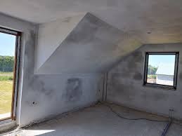 Surowe wnętrze poddasza ze świeżo nałożonym szarym tynkiem na ścianach i skośnym suficie, widoczne okna z widokiem na zieleń, na podłodze przewody.