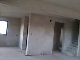 Surowe wnętrze w trakcie prac wykończeniowych: ściany pokryte szarym tynkiem, widoczne puszki elektryczne, betonowe schody w tle, naturalne światło wpadające przez okno.