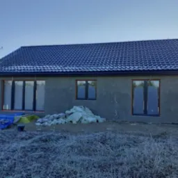 Surowy budynek jednorodzinny z ciemną dachówką i szarym tynkiem zewnętrznym, widoczne okna z ciemnymi ramami, teren wokół budynku pokryty szronem.