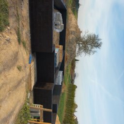 MTM DOMY SZKIELETOWE SPÓŁKA Z OGRANICZONĄ ODPOWIEDZIALNOŚCIĄ - Wznoszenie fundamentów z bloczków betonowych, zabezpieczonych hydroizolacją. Widoczne palety z materiałami budowlanymi i przygotowany teren pod budowę.