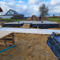 MTM DOMY SZKIELETOWE SPÓŁKA Z OGRANICZONĄ ODPOWIEDZIALNOŚCIĄ - Budowa fundamentów pod dom szkieletowy: izolacja termiczna i przeciwwilgociowa, pracownicy w tle, narzędzia budowlane. Widok na budynek w tle.