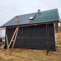 MTM DOMY SZKIELETOWE SPÓŁKA Z OGRANICZONĄ ODPOWIEDZIALNOŚCIĄ - Budowa domu szkieletowego: czarna membrana na ścianach, rusztowania, częściowo ułożone dachówki w kolorze grafitowym i wstępne elementy dachu z listew.