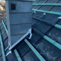 MTM DOMY SZKIELETOWE SPÓŁKA Z OGRANICZONĄ ODPOWIEDZIALNOŚCIĄ - Wzniesiony komin z szarego bloczka betonu komórkowego na tle dachu z membraną i łatami. Widok na fragment dachu i niebieskie niebo.