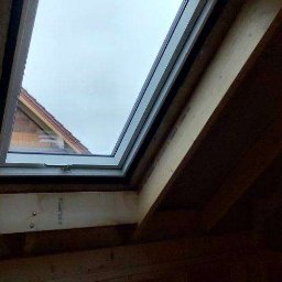 MTM DOMY SZKIELETOWE SPÓŁKA Z OGRANICZONĄ ODPOWIEDZIALNOŚCIĄ - Montaż okna dachowego w drewnianej konstrukcji dachu. Widok z wnętrza poddasza na okno z szybą, przez którą widać fragment dachu sąsiedniego budynku.