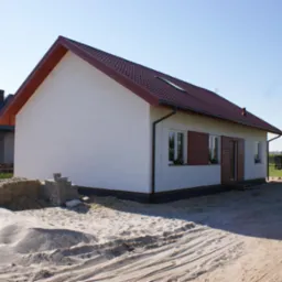 Niski, parterowy budynek z białymi ścianami i brązowym dachem, widoczny z boku, z stertą piasku i bloczków fundamentowych na pierwszym planie, sugerujący etap budowy.