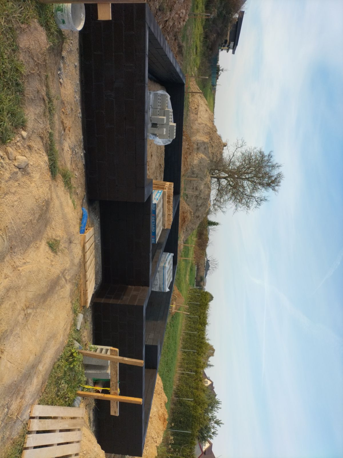 Wznoszenie fundamentów z bloczków betonowych, zabezpieczonych hydroizolacją, na budowie domu. Widoczne materiały budowlane i otaczający teren.