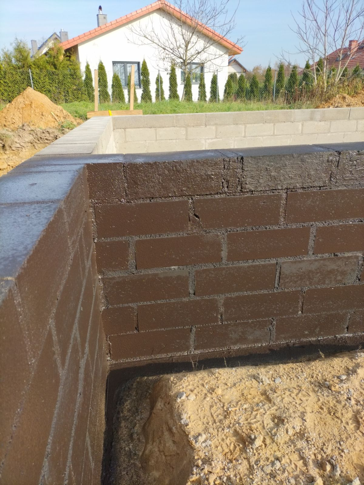 Hydroizolacja fundamentów z cegły w Twardogórze, zabezpieczona brązową powłoką. W tle widoczny dom w trakcie budowy i pryzma piasku.