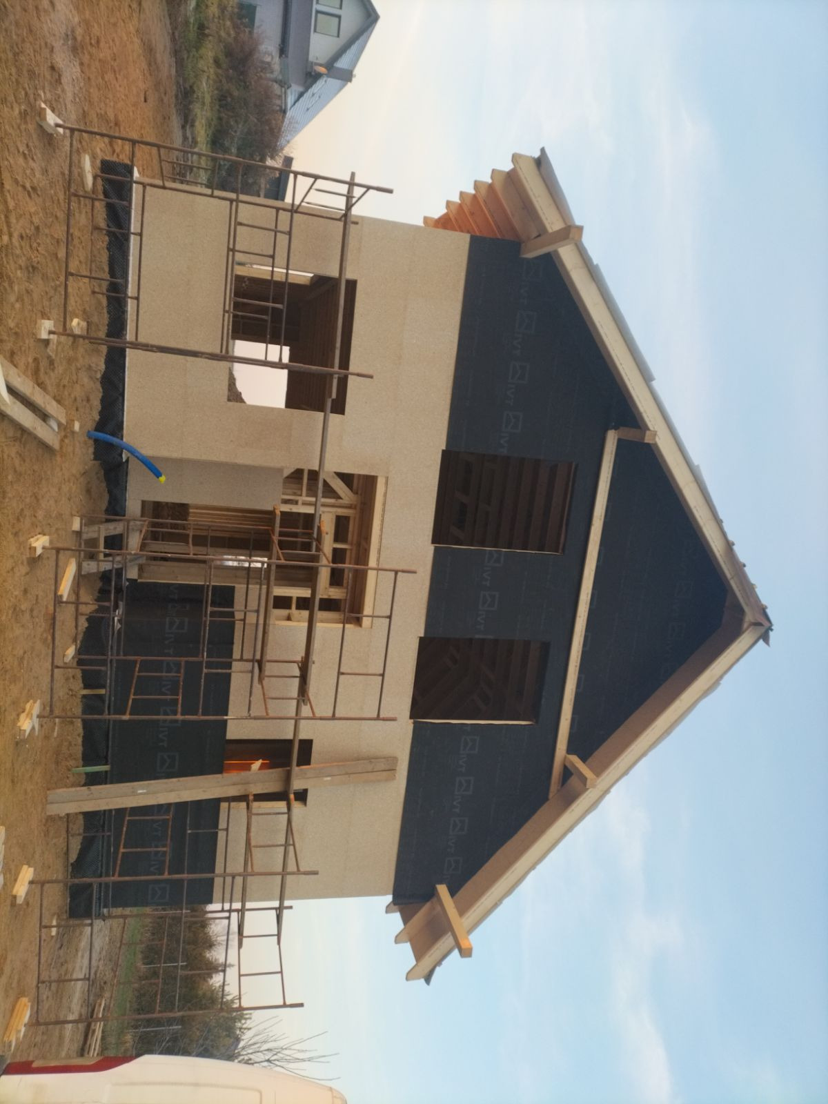 Budowa domu szkieletowego w trakcie: widoczne ściany z otworami okiennymi, rusztowania i fragment dachu z membraną. Ujęcie z dołu, niebo w tle.