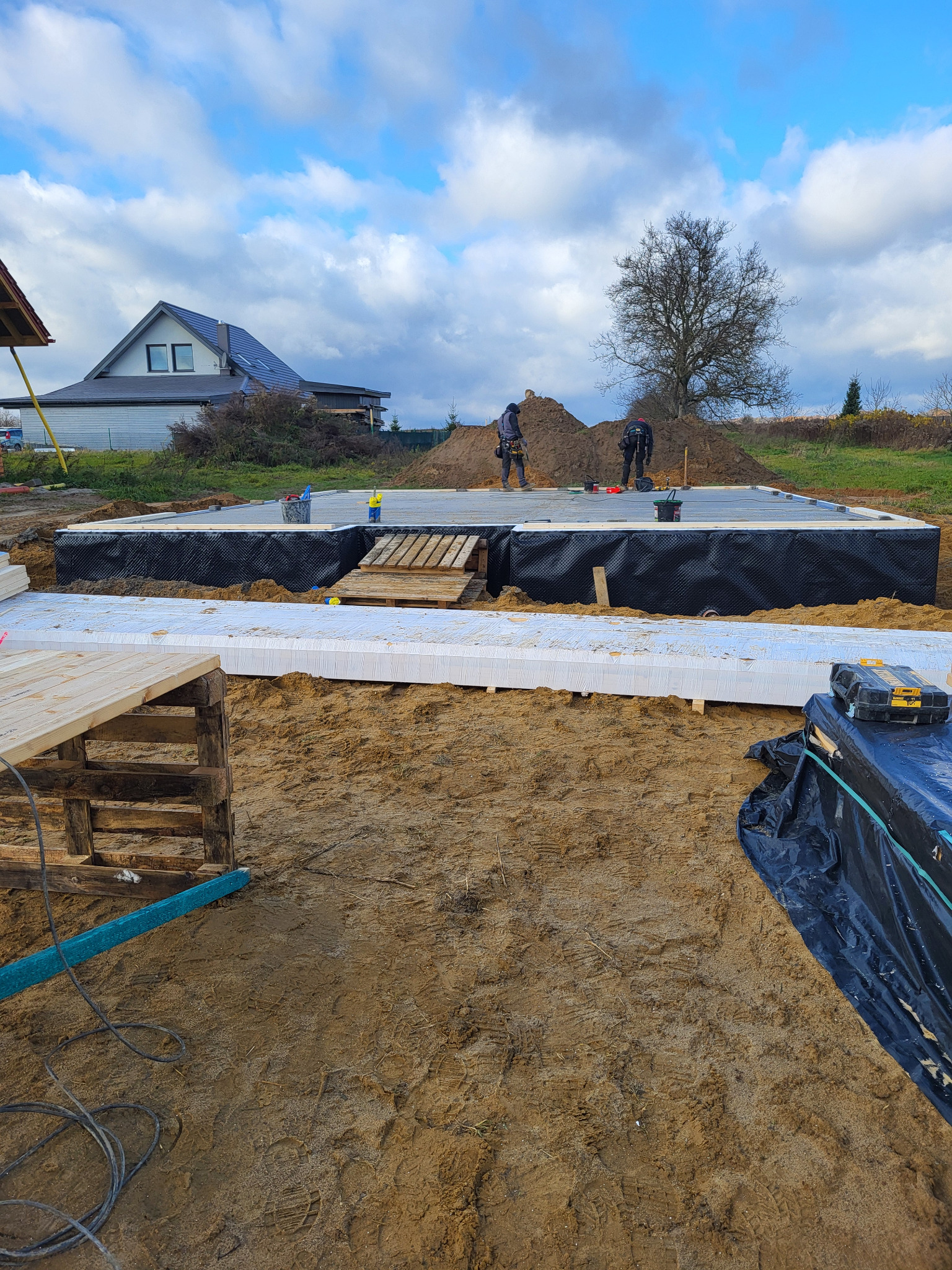 Budowa fundamentów pod dom szkieletowy: izolacja termiczna i przeciwwilgociowa, pracownicy w tle, narzędzia budowlane. Widok na budynek w tle.