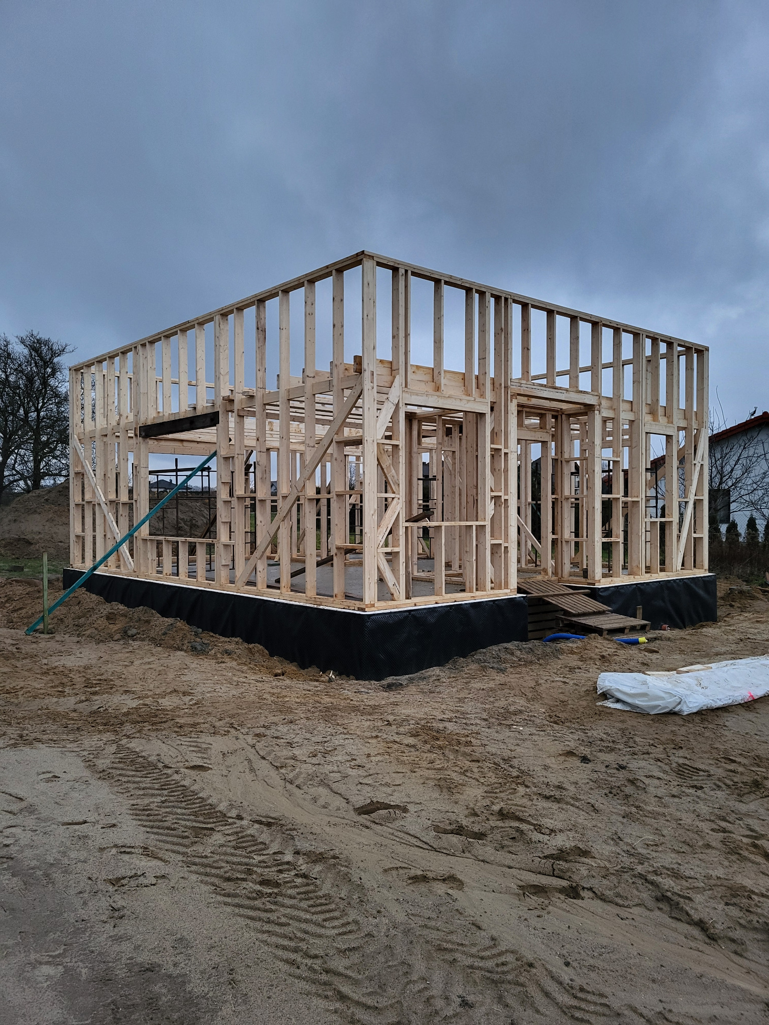 Konstrukcja szkieletowa domu w trakcie budowy, widoczne drewniane elementy, fundament zabezpieczony czarną folią, pochmurne niebo w tle.