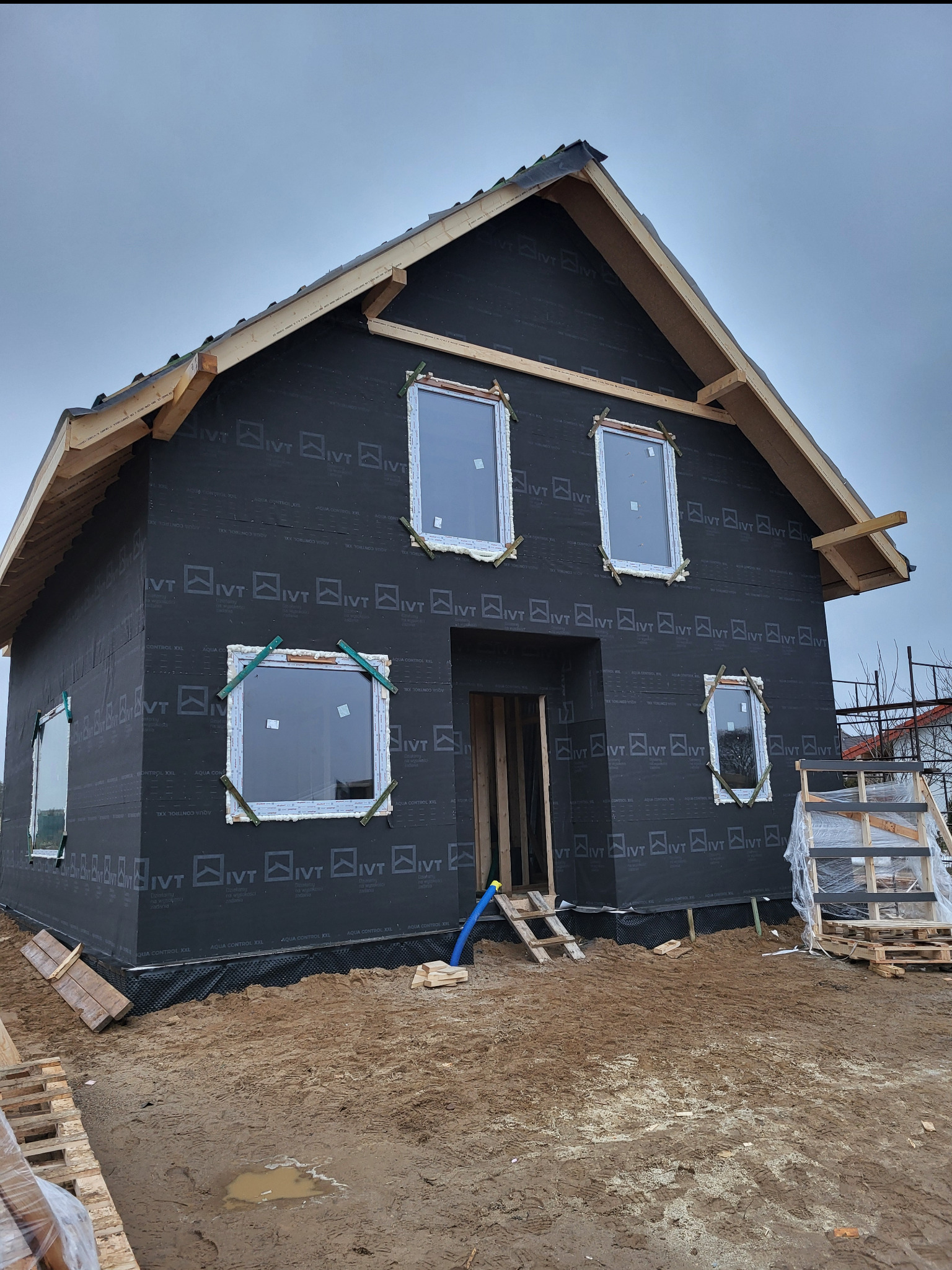 Surowy dom szkieletowy z czarną membraną, widoczne okna i drewniana konstrukcja dachu w trakcie budowy. Palety i rusztowania w otoczeniu.