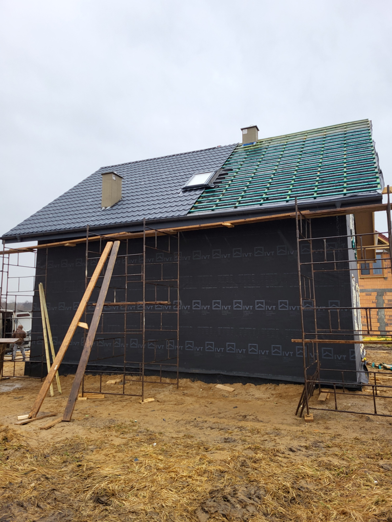 Budowa domu szkieletowego: czarna membrana na ścianach, rusztowania, częściowo ułożone dachówki w kolorze grafitowym i wstępne elementy dachu z listew.