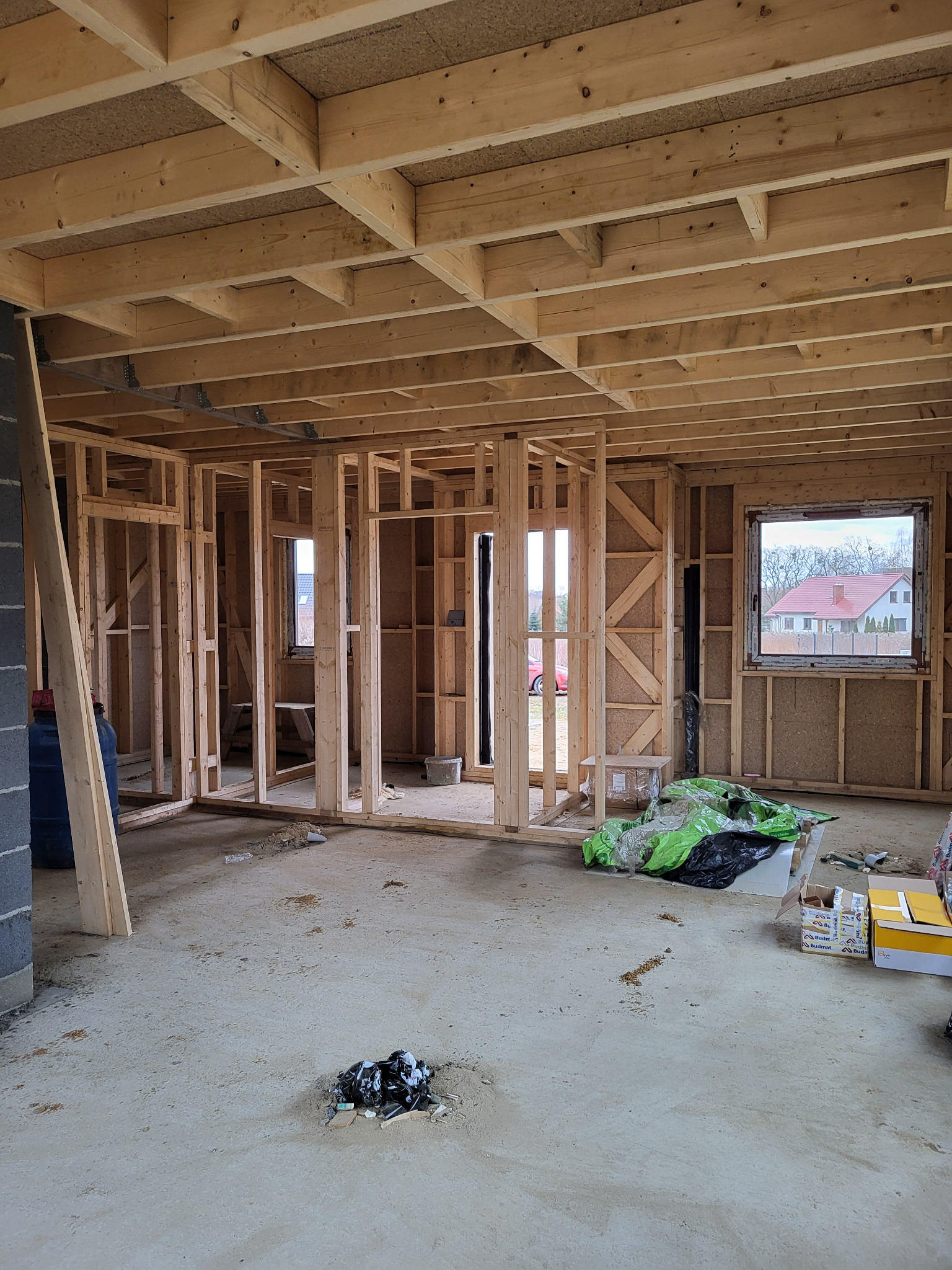 Wnętrze w trakcie budowy domu w technologii szkieletowej. Widoczne drewniane elementy konstrukcyjne ścian, stropu i otwory okienne. Na podłodze materiały budowlane i śmieci.