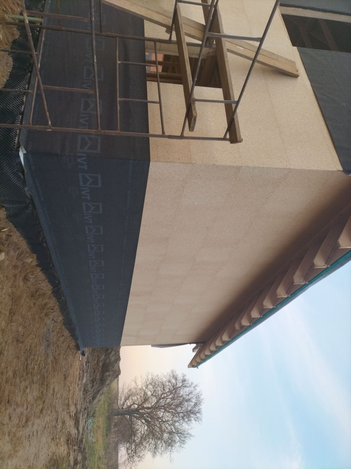 Budowa domu w konstrukcji szkieletowej: widok na ścianę z płyt OSB, zabezpieczoną czarną membraną i rusztowaniem. W tle drzewo bez liści.