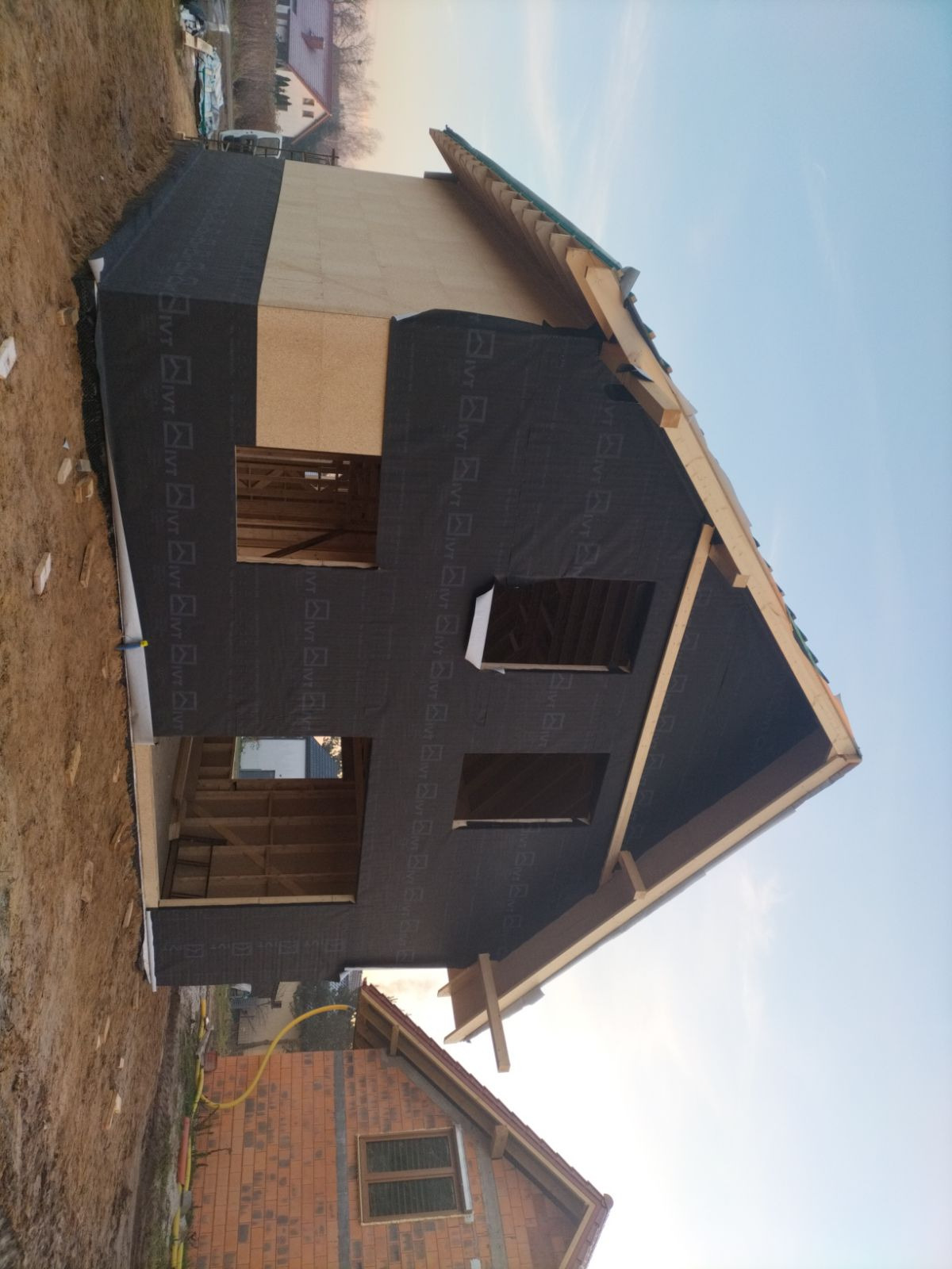 Konstrukcja domu szkieletowego w trakcie budowy: widoczne drewniane elementy konstrukcyjne, ściany pokryte membraną. Częściowo ocieplony budynek z widocznymi otworami okiennymi.