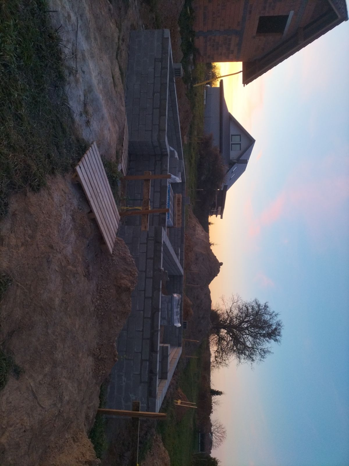 Wznoszenie fundamentów z bloczków betonowych pod budowę domu na tle zachodzącego słońca. Widoczne drewniane podpory i częściowo zarys budynku w tle.