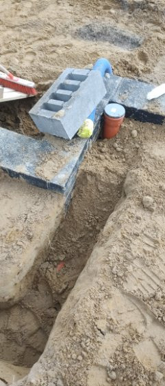 Wykop w ziemi z elementami konstrukcyjnymi, fragmentem rury i betonowym bloczkiem. Widoczne ślady prac ziemnych i przygotowanie do dalszych etapów budowy.