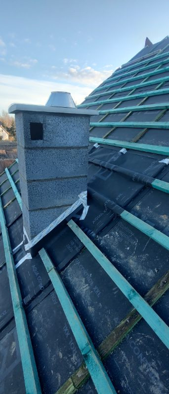 Wzniesiony komin z szarego bloczka betonu komórkowego na tle dachu z membraną i łatami. Widok na fragment dachu i niebieskie niebo.