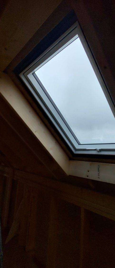 Wewnętrzny widok okna dachowego z drewnianą ramą, zamontowanego w konstrukcji dachu o drewnianym wykończeniu, z widokiem na pochmurne niebo.