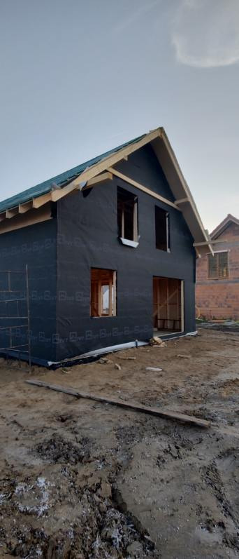 Budowa domu szkieletowego: widok na dwukondygnacyjny budynek z czarną membraną na ścianach i częściowo wykonanym dachem. W tle widoczny sąsiedni dom z czerwonej cegły.