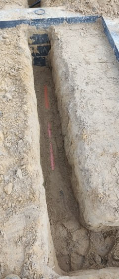 Wykop w ziemi z widocznymi pomarańczowymi rurami instalacyjnymi. Wykop zabezpieczony metalową konstrukcją. Widok z góry na wykop.