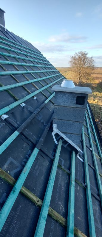 Dach z membraną i łatami pod dachówkę, komin z szarego pustaka, widok z dołu na tle pola i drzewa, w słoneczny dzień.