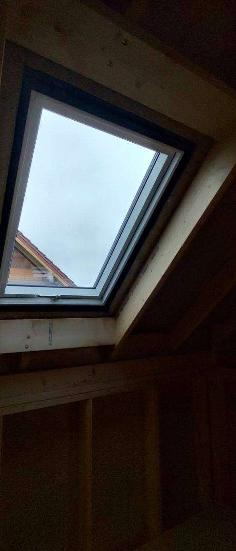 Montaż okna dachowego w drewnianej konstrukcji dachu. Widok z wnętrza poddasza na okno z szybą, przez którą widać fragment dachu sąsiedniego budynku.