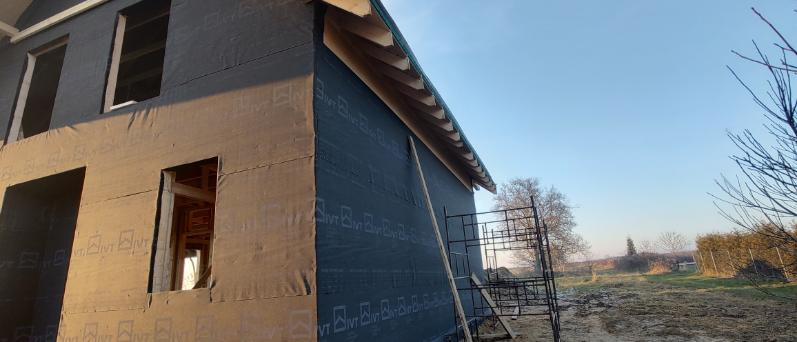 Budowa domu w technologii szkieletowej: ściany z membraną i wycięte otwory okienne, widoczne rusztowanie, drzewa i błękitne niebo w tle.