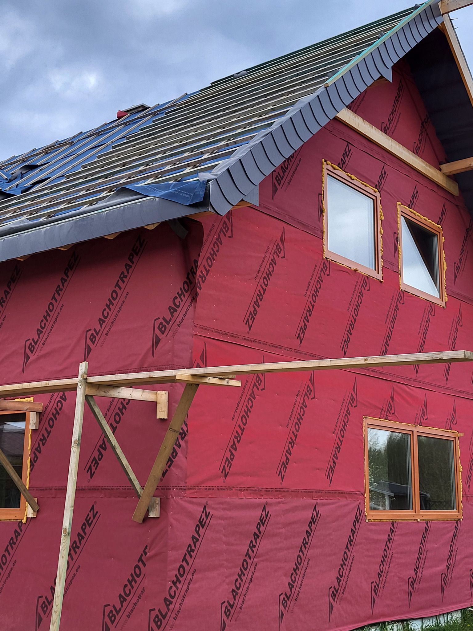 Wznoszenie domu szkieletowego: widoczne drewniane elementy konstrukcji dachu, ściany osłonięte czerwoną membraną z nadrukiem, okna z brązowymi ramami.