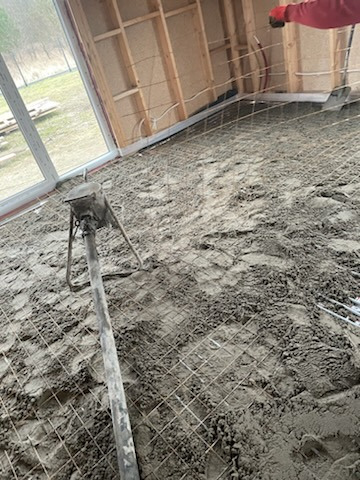 Wnętrze w trakcie wylewki podłogi z siatką zbrojeniową i maszyną do wylewania betonu. Widoczne drewniane elementy konstrukcji szkieletowej.