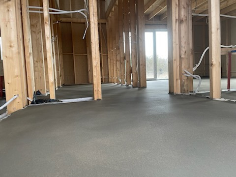 Wnętrze w trakcie budowy: surowy stan z drewnianą konstrukcją ścian i wylaną, świeżą posadzką. Instalacje elektryczne widoczne wzdłuż ścian.