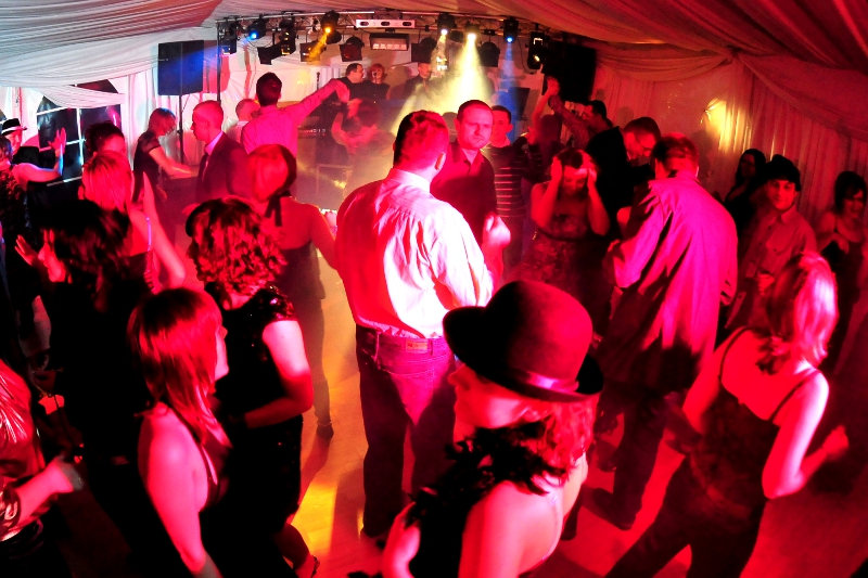 Tłum ludzi tańczących na parkiecie w czerwonym oświetleniu dyskotekowym, w tle widoczna scena z występującymi muzykami.