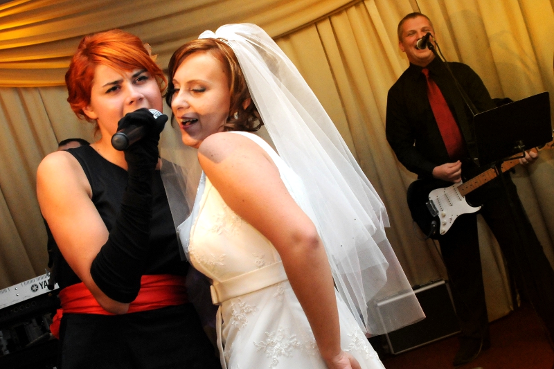Wokalistka z mikrofonem w długich rękawiczkach i czerwonym pasem, śpiewa obok panny młodej w welonie; w tle gitarzysta z laptopem na statywie.