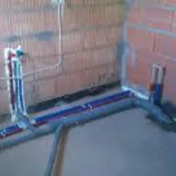 Instalacja wodno-kanalizacyjna w surowym wnętrzu: rury doprowadzające wodę w kolorze niebieskim i czerwonym, szare rury odpływowe, widoczne przewody elektryczne.