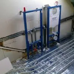 Białe rury ogrzewania podłogowego ułożone na siatce, z widoczną instalacją hydrauliczną w tle. Ujęcie z góry pod kątem, ukazujące etap montażu.