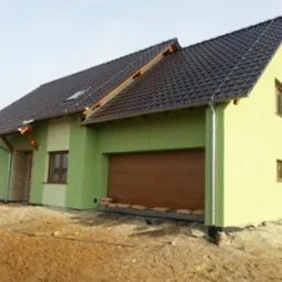 Zakończony dom z zieloną elewacją, ciemną dachówką i drewnianą bramą garażową. Widoczne elementy wykończeniowe z kamienia przy wejściu i teren wokół domu w stanie surowym.