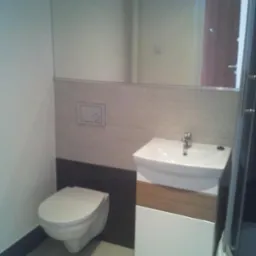 Nowoczesna łazienka po remoncie: podwieszana toaleta, umywalka z szafką i duże lustro odbijające fragment drzwi wejściowych. Minimalistyczny design, jasne płytki.