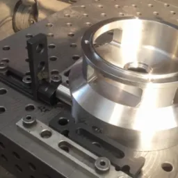 Metalowy element obrabiany na perforowanym stole spawalniczym z użyciem czarnych uchwytów i narzędzi, widoczne ślady obróbki, oświetlenie warsztatowe.