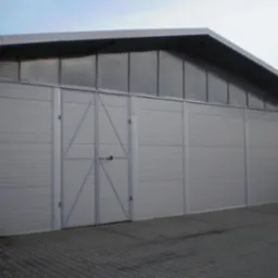Gotowa hala magazynowa z szarej blachy trapezowej, z dwuskrzydłowymi drzwiami na środku, widok z zewnątrz na kostce brukowej.