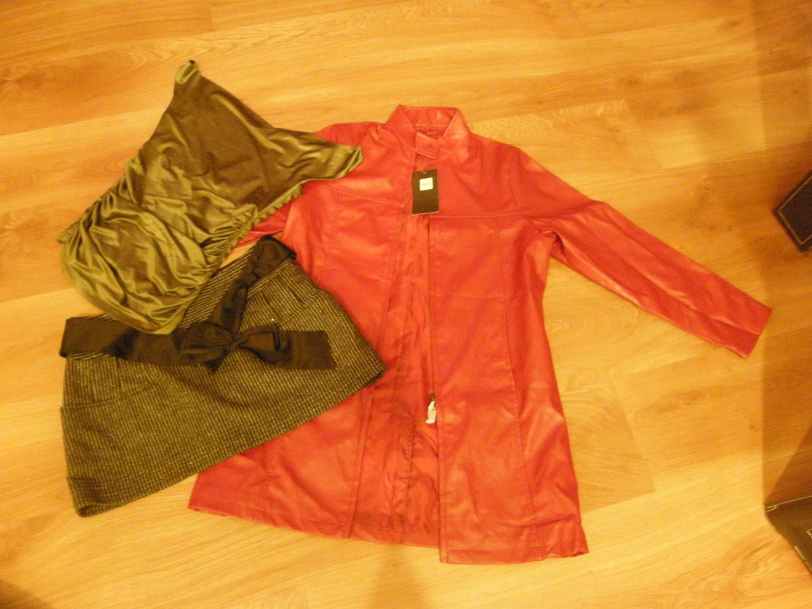 Rozłożona na drewnianej podłodze czerwona kurtka z metką, oliwkowa drapowana bluzka i krótka tweedowa spódnica z czarną kokardą.