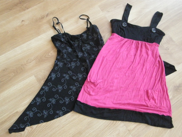 Dwie sukienki na ramiączkach leżące na drewnianej podłodze: czarna z delikatnym wzorem i różowo-czarna z kieszeniami i ozdobnymi guzikami.