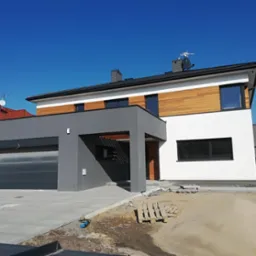 Nowoczesny dom jednorodzinny z garażem w bryle, wykończony drewnem i białym tynkiem, widoczny fragment podjazdu i ogród w trakcie prac wykończeniowych.