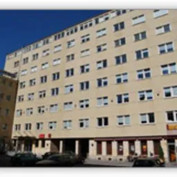 Wielopiętrowy budynek z zamontowanymi oknami PCV, widoczny z dołu w słoneczny dzień, z zaparkowanymi samochodami i lokalami usługowymi na parterze.