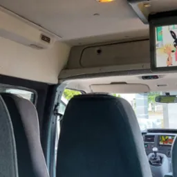 Wnętrze busa z zamontowanym monitorem sufitowym wyświetlającym bajkę dla dzieci, widok z tylnych siedzeń na deskę rozdzielczą i przednią szybę.